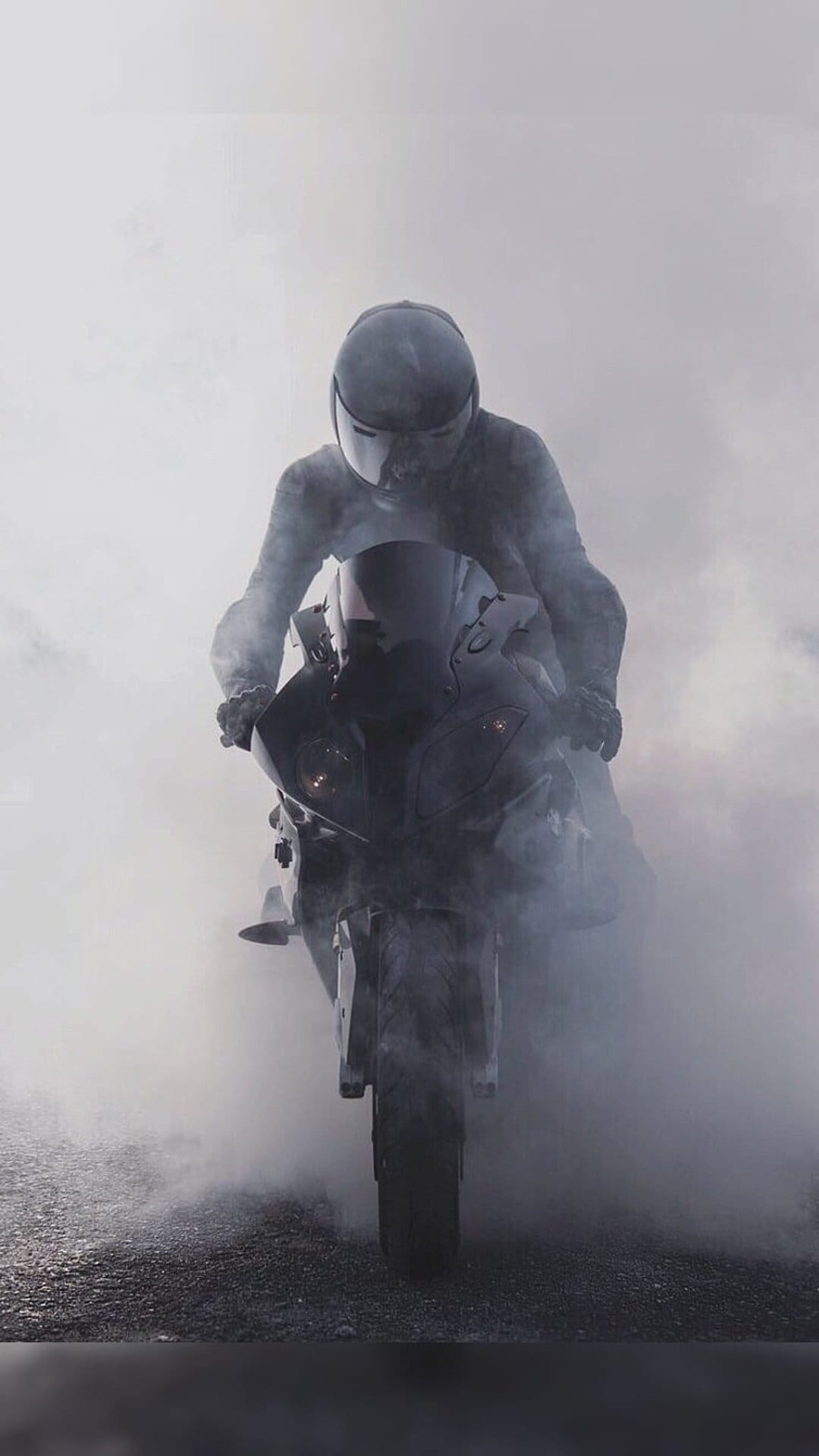 Motorcyclist in black gear rides through dense white smoke on a dark sportbike with glowing headlights on gravel.