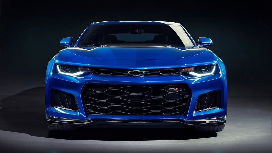 Front view of a blue Chevrolet Camaro ZL1 with glowing LED headlights and a black honeycomb grille in a dark studio.