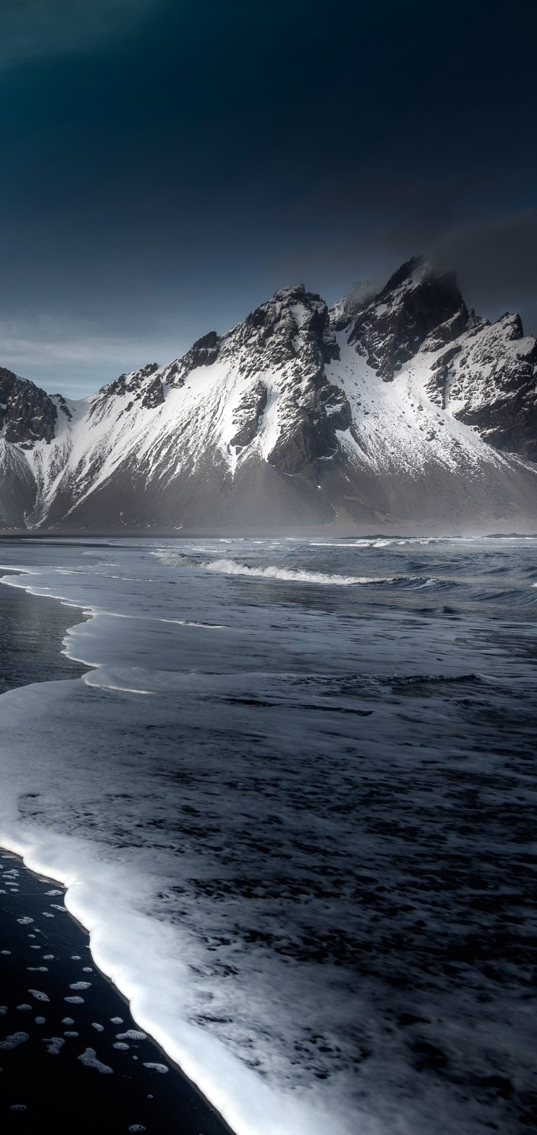 Black sand beach with white foam waves meeting snowy mountains under a dark, moody sky in vertical orientation.