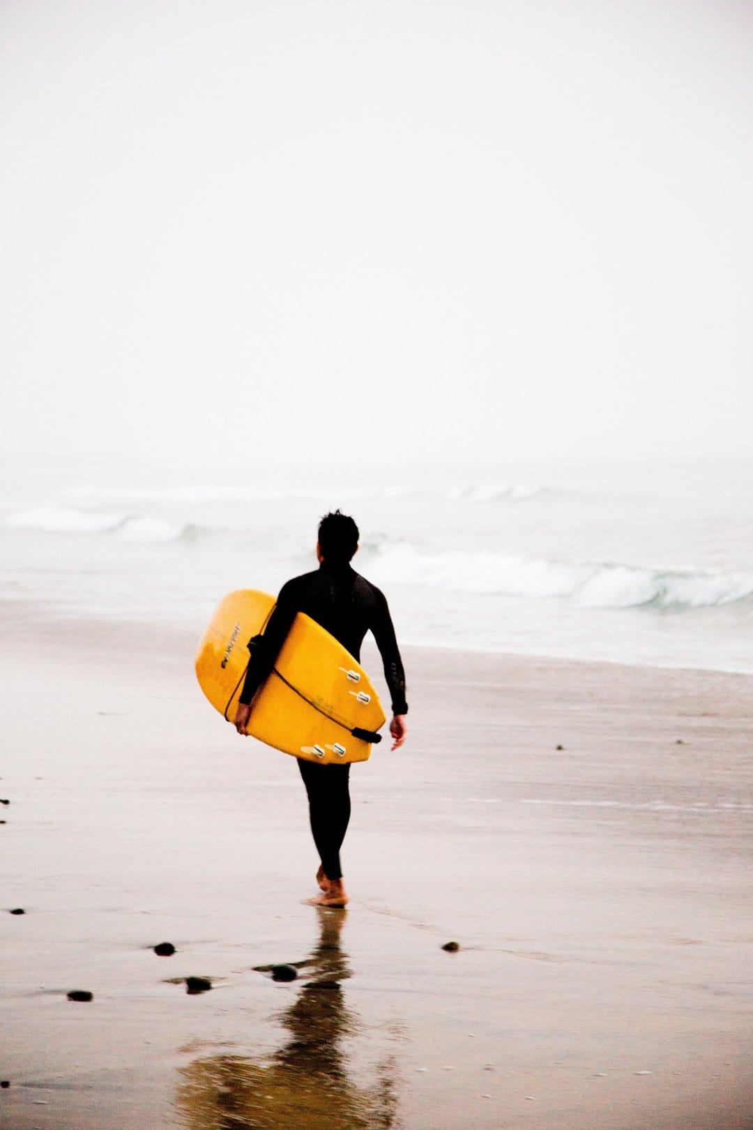 Surfer in a black wetsuit carries a yellow board on a misty beach with reflections visible in the wet sand.