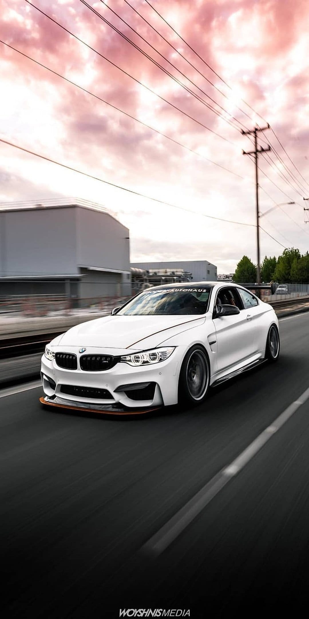 White BMW M4 coupe with custom body kit cruising an urban road beneath a pink and orange cloudy sunset sky.