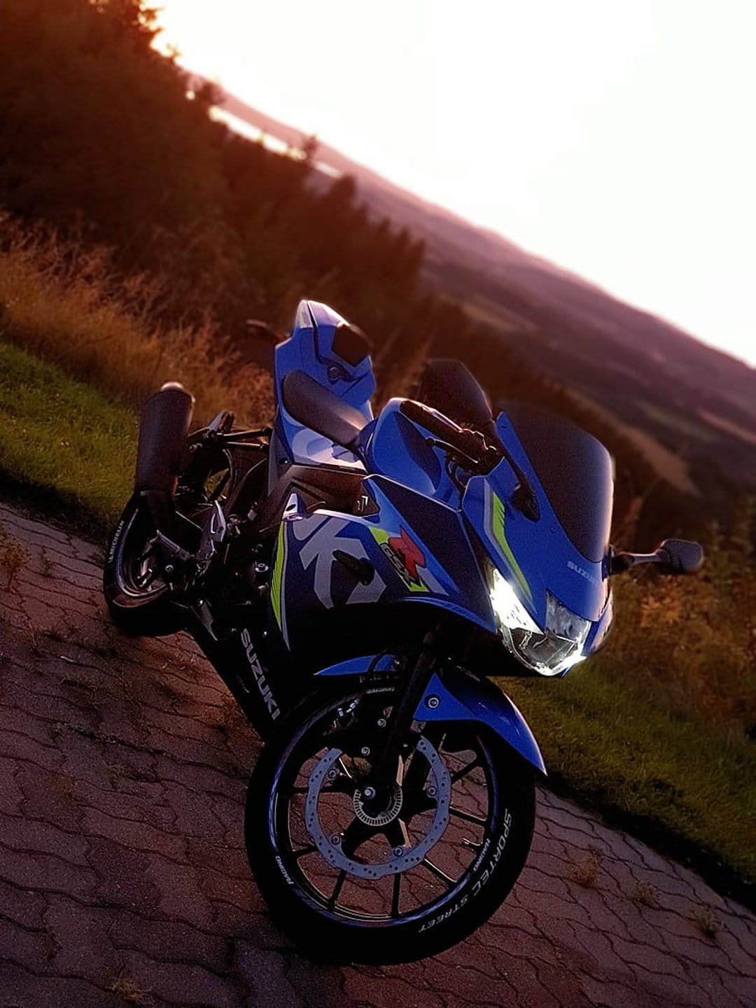 Blue Suzuki sportbike with headlights on parked on a hillside road overlooking a scenic sunset and water.