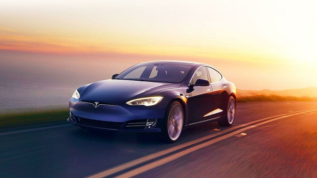 Blue Tesla Model S driving on a coastal road at sunset with golden sky reflections on the car body.