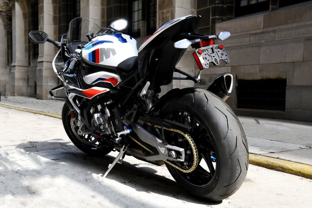Rear view of a white and blue BMW M 1000 RR motorcycle with a gold chain on a sunny stone sidewalk.