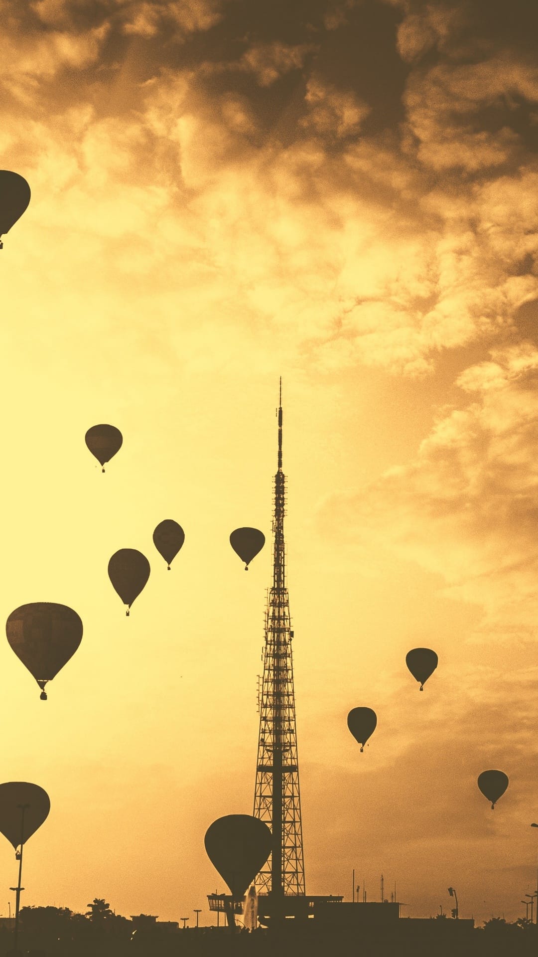 Hot air balloons ascend past a tall communication tower into a hazy golden sky during a monochromatic sunset.
