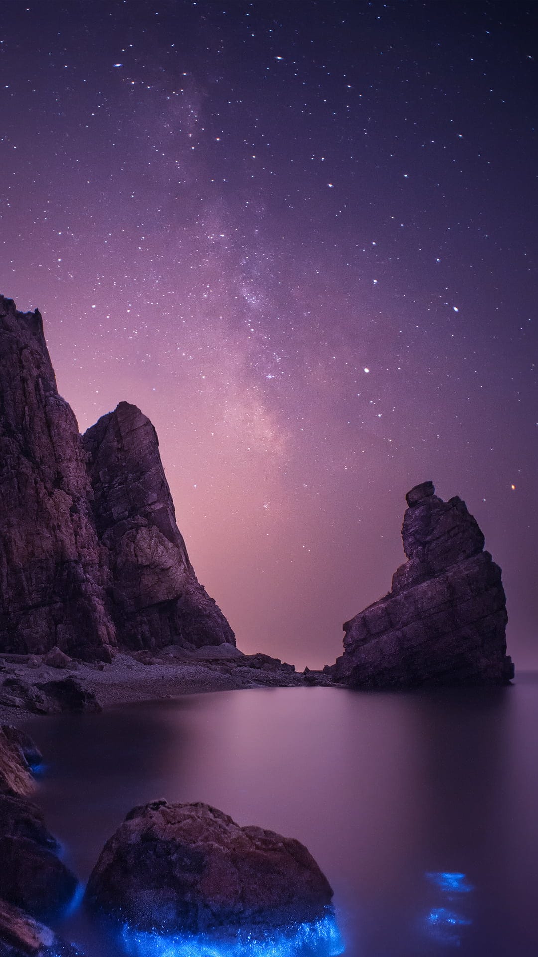 Bioluminescent blue tide glows beneath a detailed Milky Way galaxy arching over rugged seaside cliffs at night.