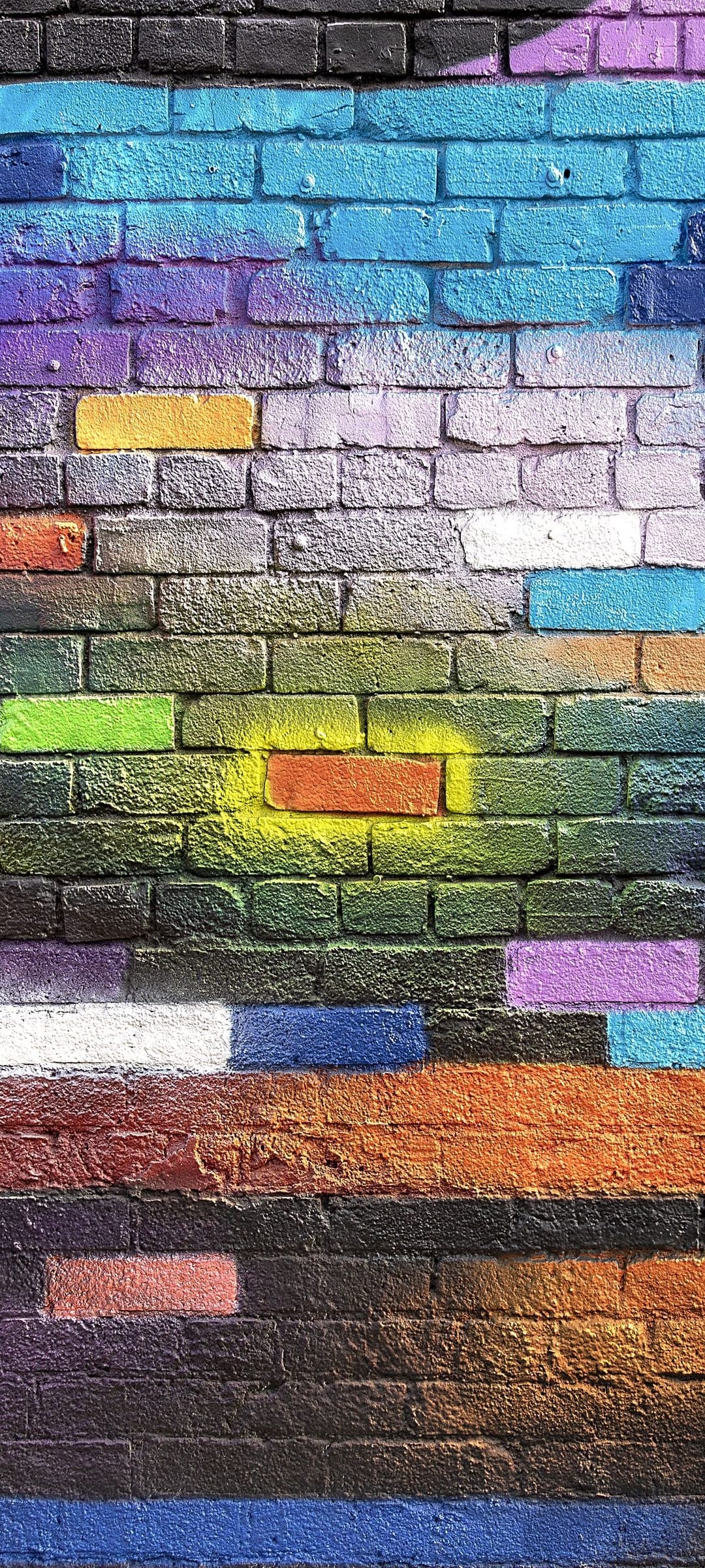 Multicolored horizontal graffiti stripes with spray paint splatters cover a textured brick wall surface.