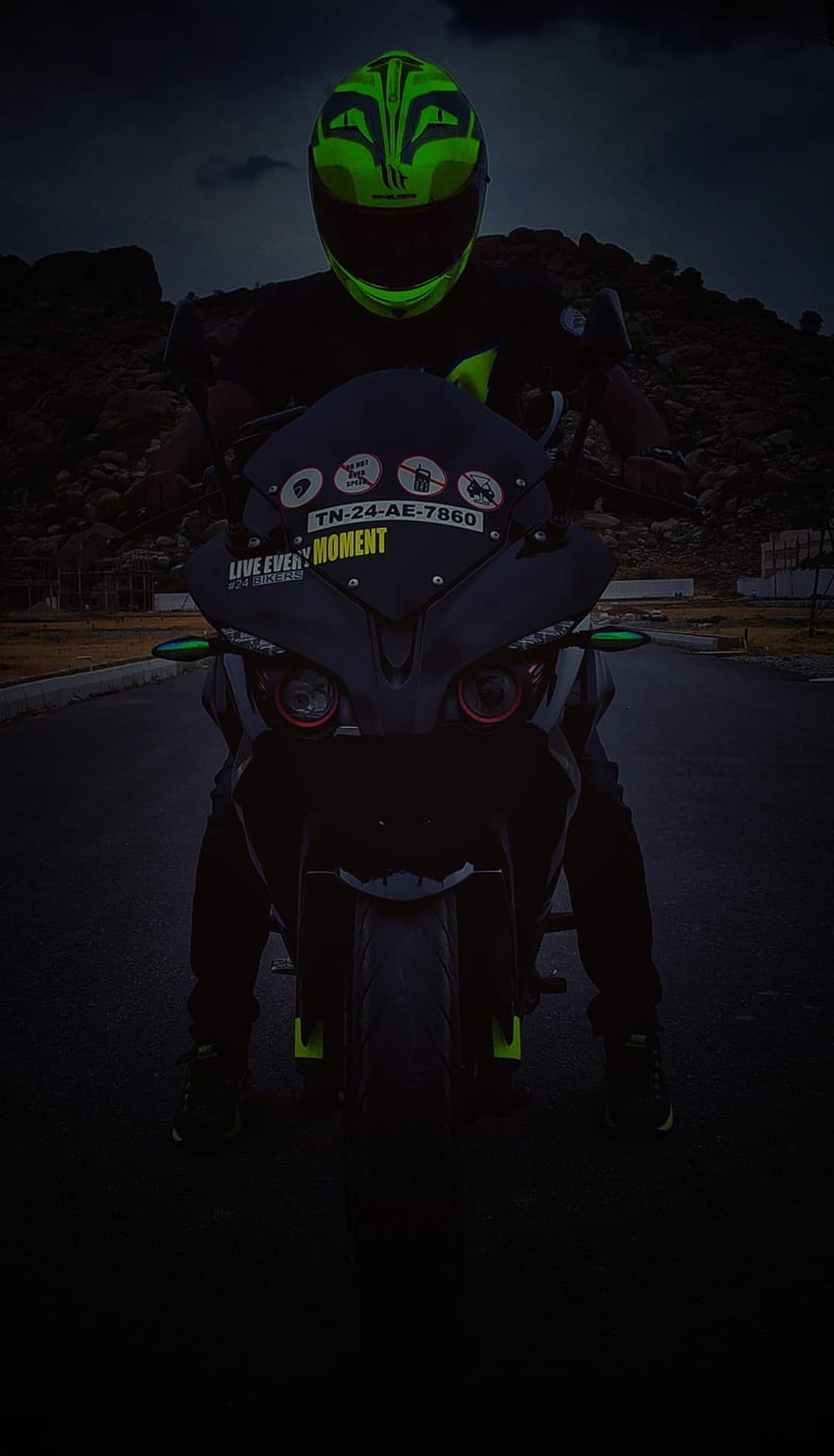 Motorcyclist in a neon green helmet sits on a black bike with red circular headlights against a rocky backdrop.