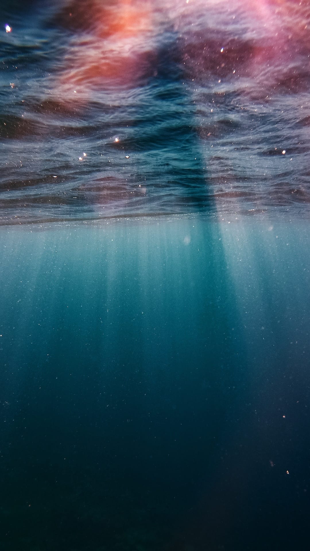 Sunlight beams pierce deep blue ocean water with floating particles and surface ripples in a vertical view.