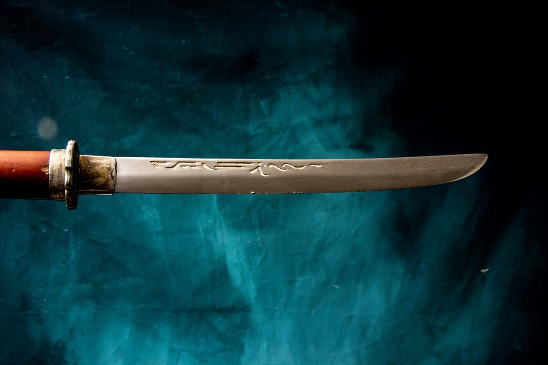 Katana blade with wave engravings and a red hilt set against a dark teal smoky background in a close-up view.