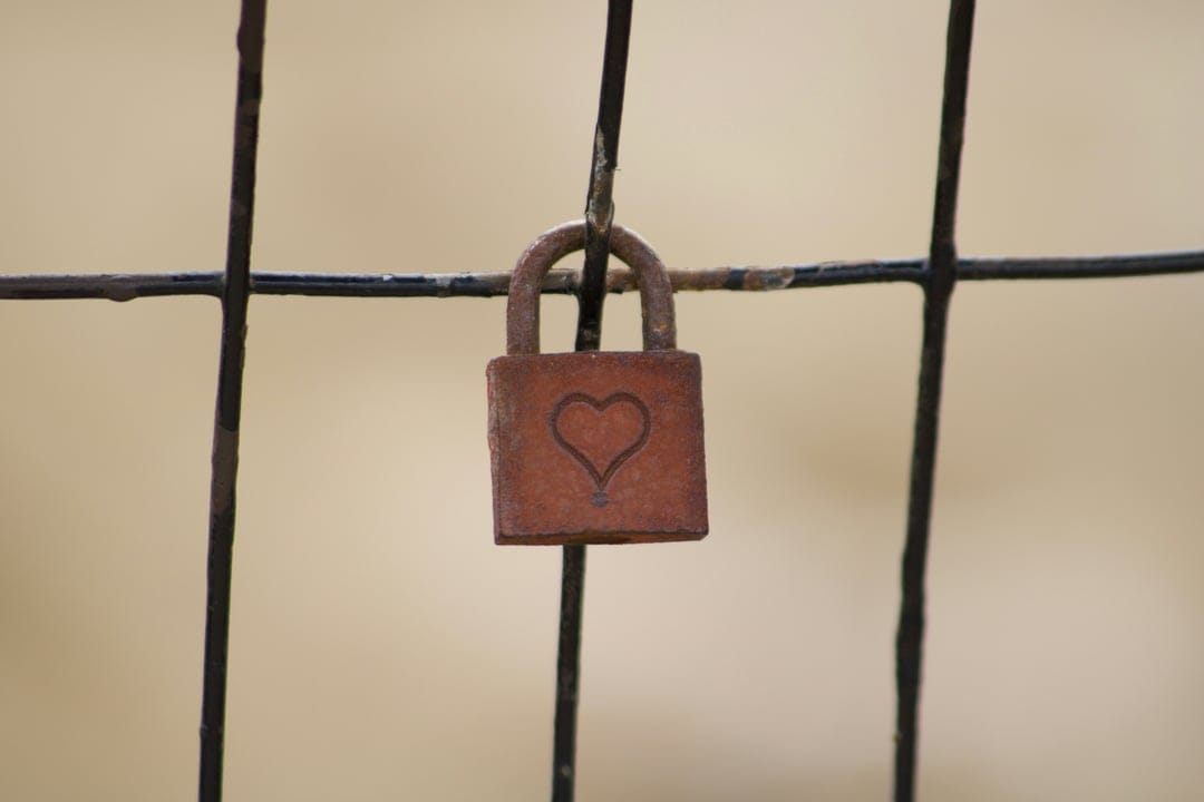 Rusty padlock with a small etched heart hangs on a dark chain-link fence against a soft-focus background.