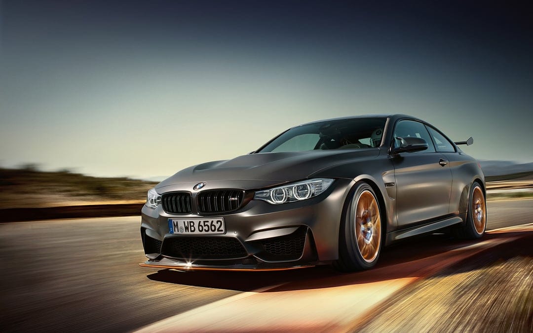 Dark grey BMW M4 GTS with orange accents racing on a track with motion blur under a clear sky.
