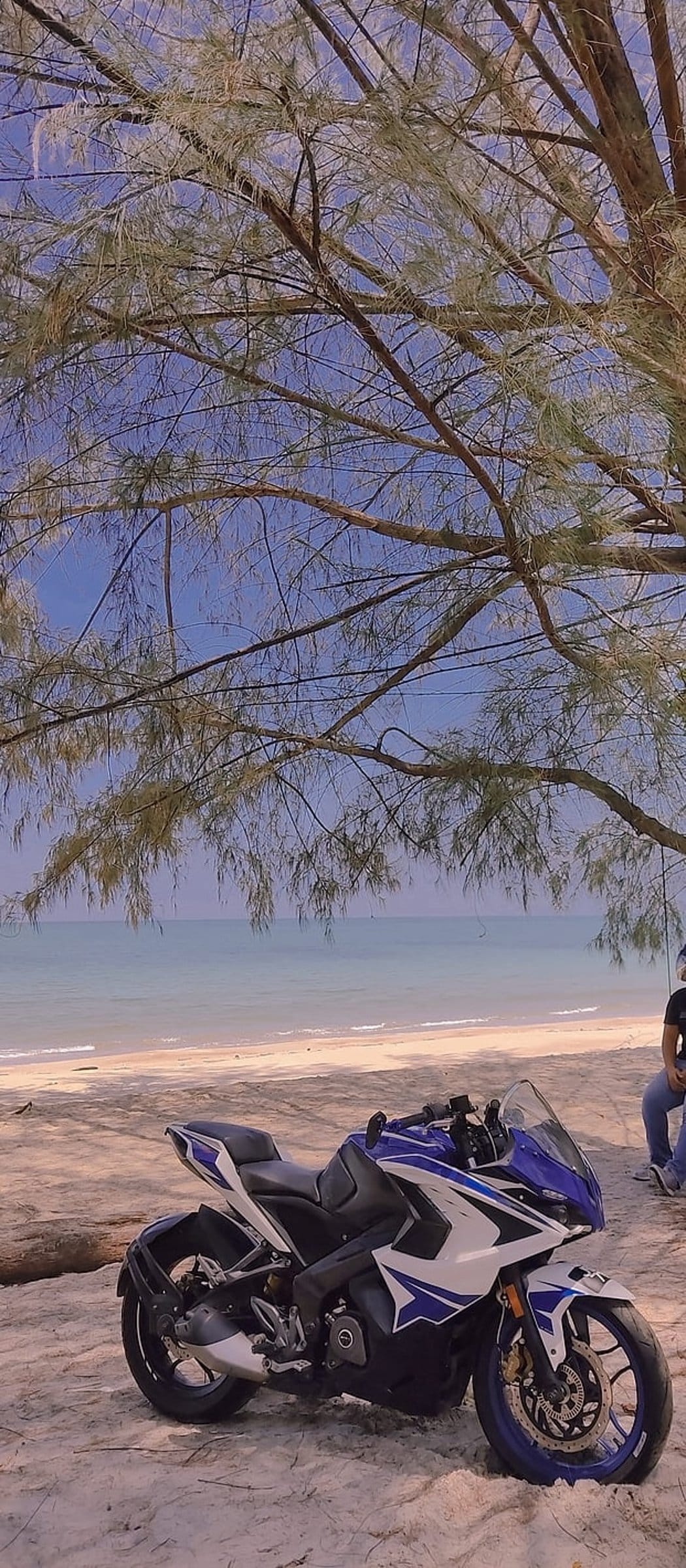 Blue and white sports motorcycle parked on a sandy beach with the ocean and a clear sky in the background.