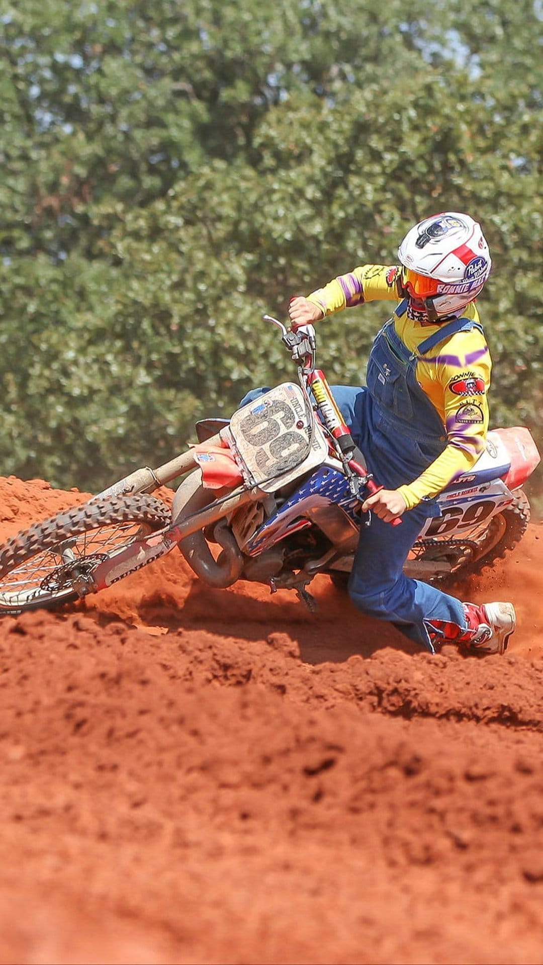 Motocross rider in blue gear leans a racing bike into a dusty turn on a red clay track during a daytime event.