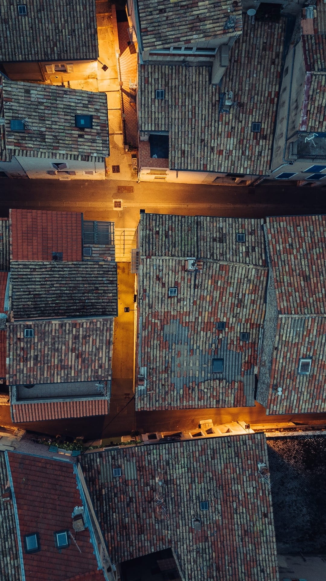 Terracotta roof tiles on old buildings glow with amber streetlights in a narrow European alleyway at twilight.