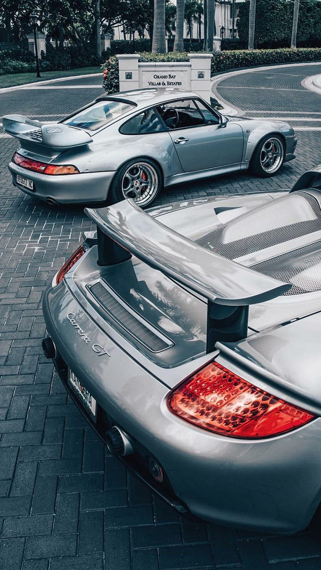 Silver Porsche Carrera GT rear wing and taillights parked on a cobblestone driveway near an elegant entrance.