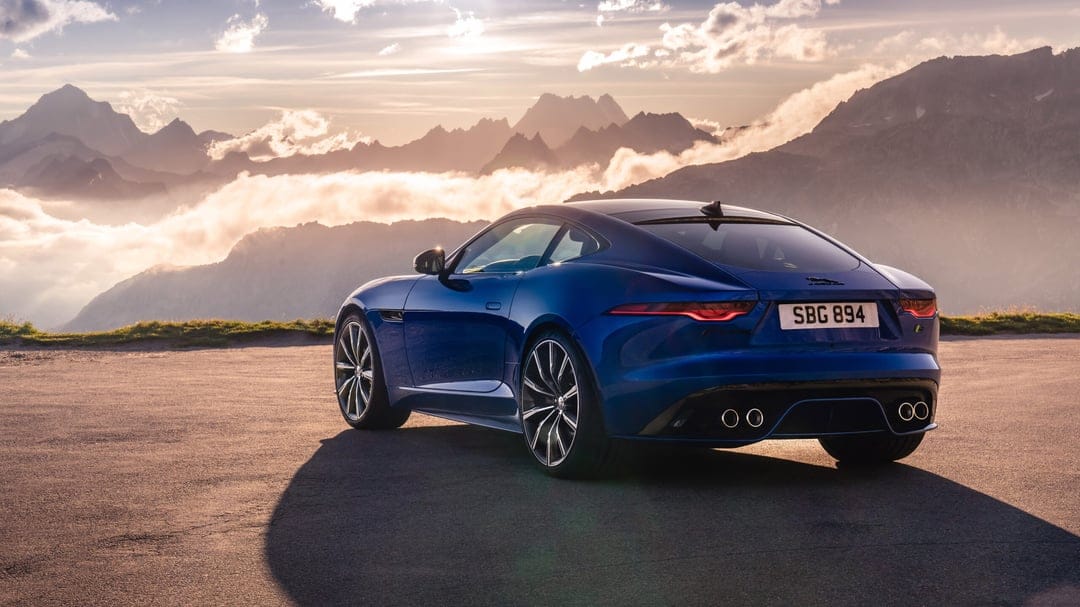 Blue sports car parked on a mountain road with sunset light hitting misty peaks and clouds in the background.