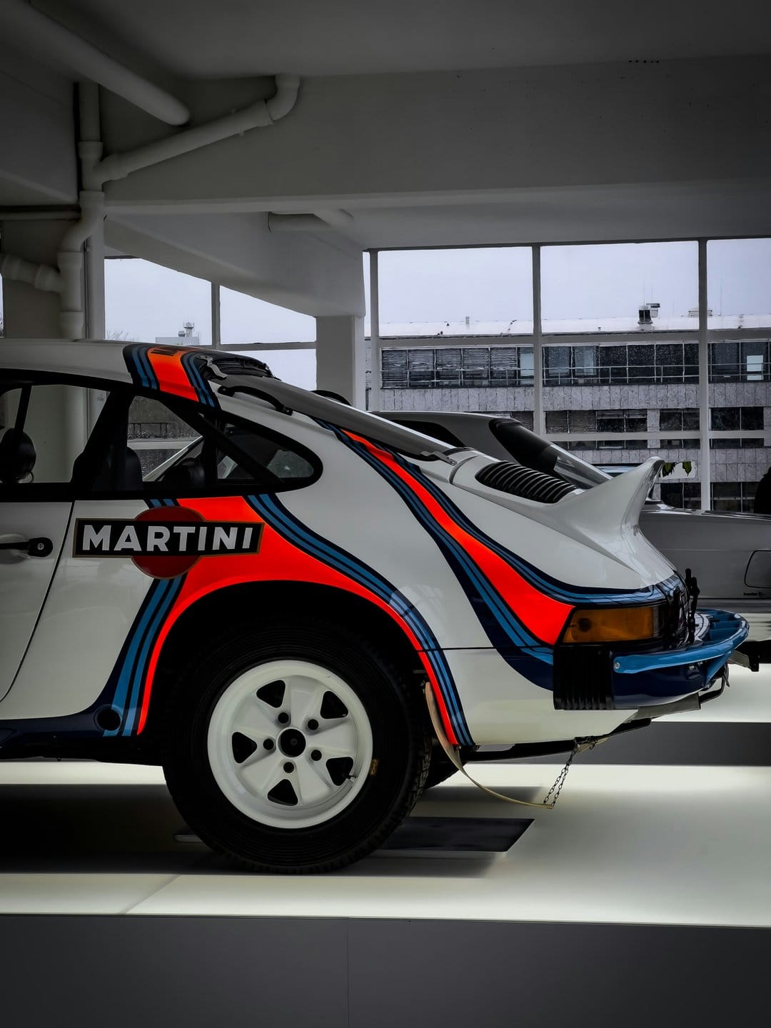Vintage Porsche 911 Turbo with Martini Racing livery stripes displayed in a brightly lit museum exhibition hall.