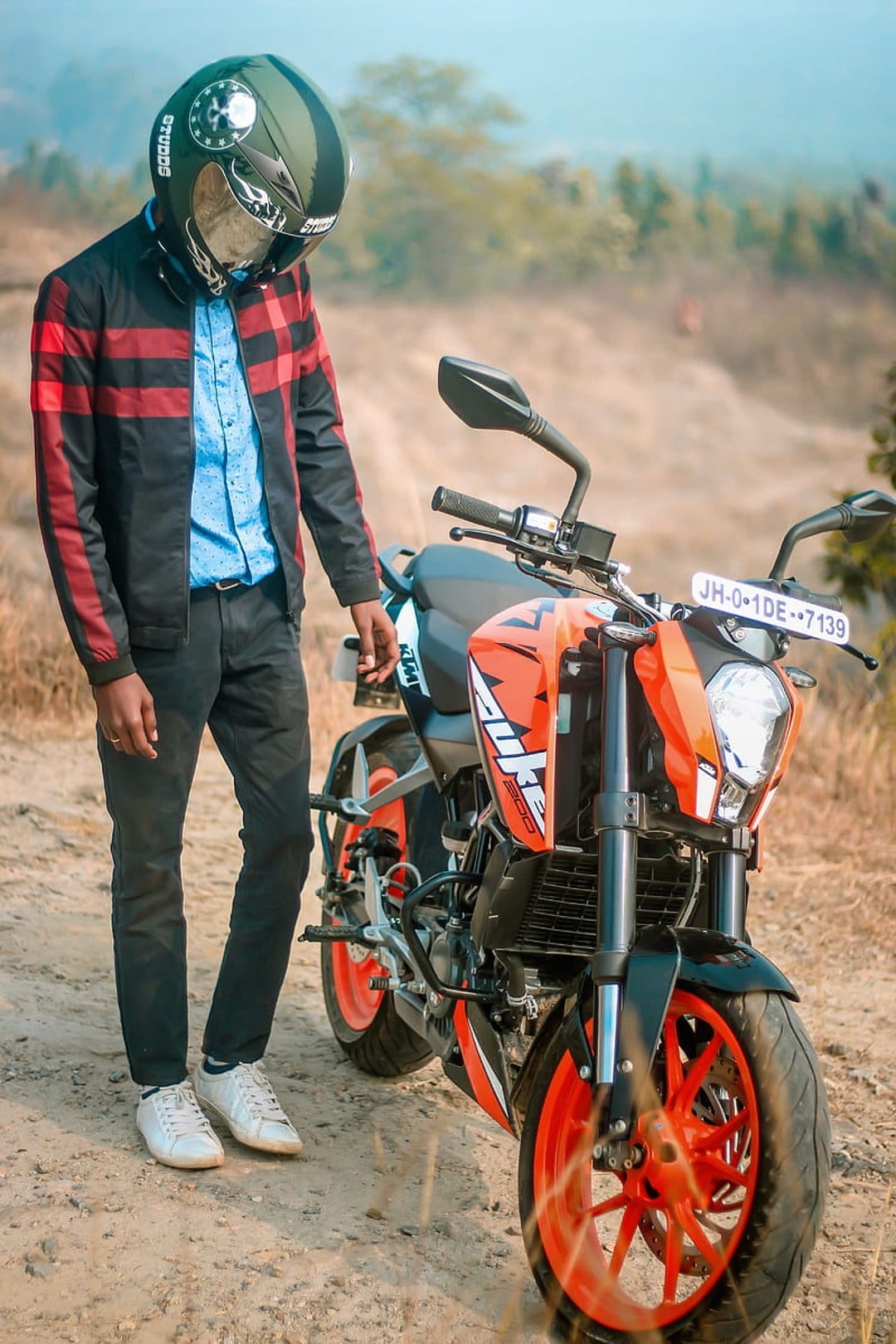 Biker in a black helmet and red jacket stands next to an orange KTM motorcycle with a blurred green background.