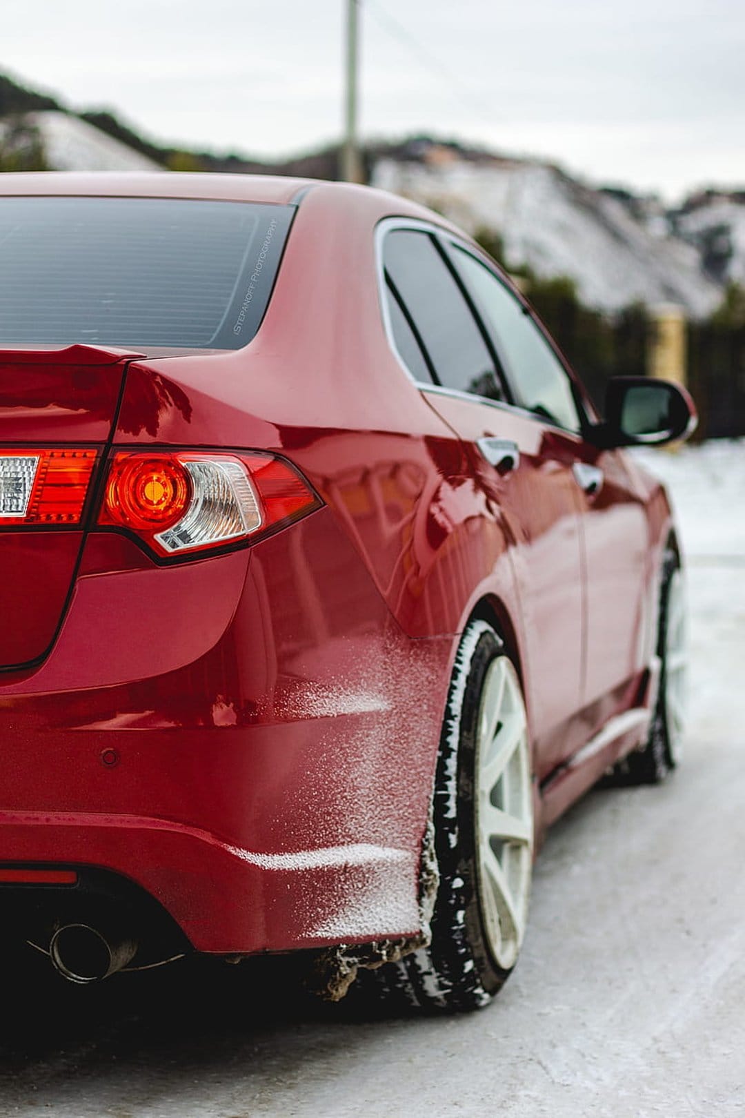 Red sedan rear panel with white wheels parked on a snowy ground against a blurred winter landscape background.