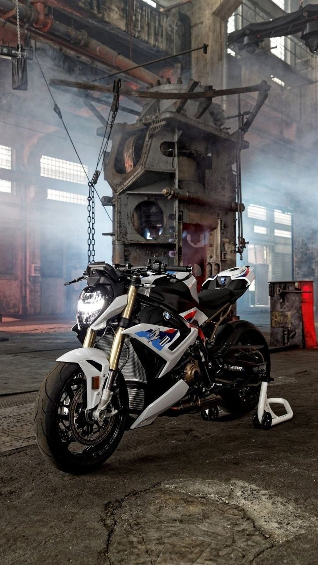 BMW S1000R motorcycle in a white finish stands amidst thick smoke and steel chains inside a dark warehouse.