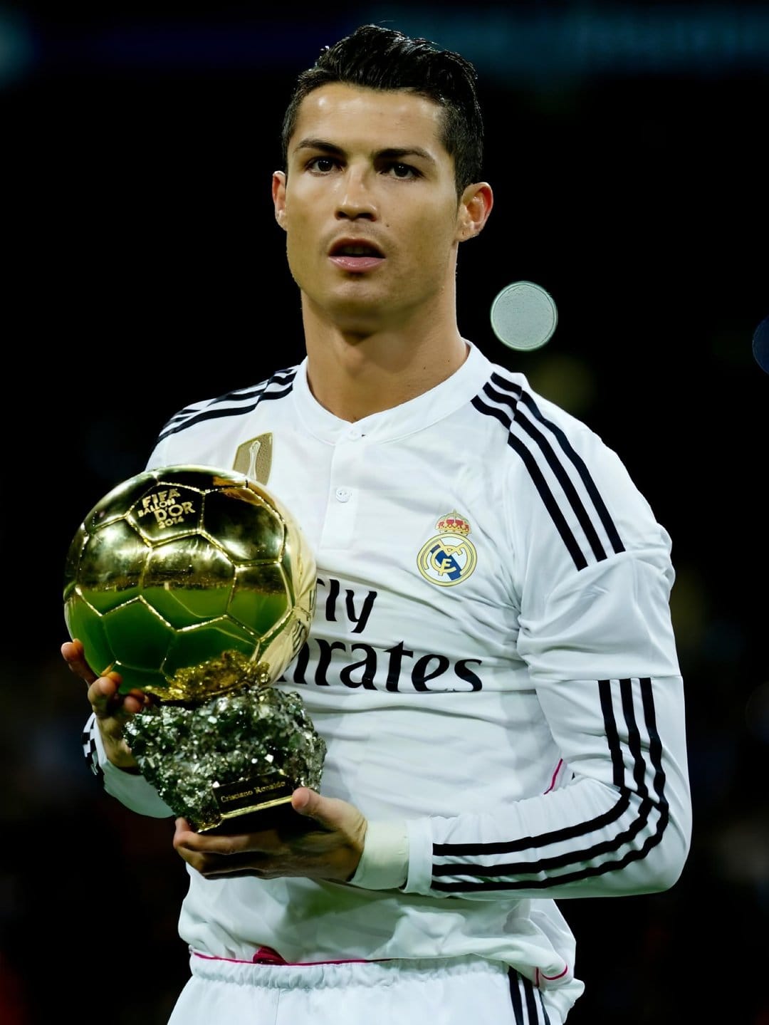 Cristiano Ronaldo holds a gold Ballon d'Or trophy while wearing a white Real Madrid kit in a stadium setting.