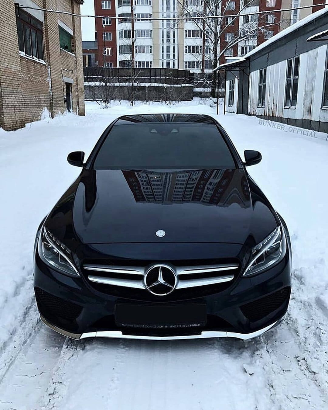 Black Mercedes-Benz C-Class sedan parked on a snowy urban street with building reflections on its polished hood.