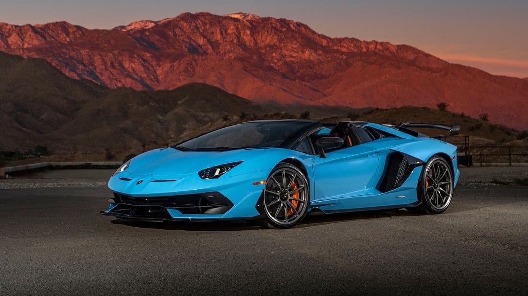 Blue Lamborghini Aventador SVJ Roadster parked on asphalt with sunset-lit mountains in the background.