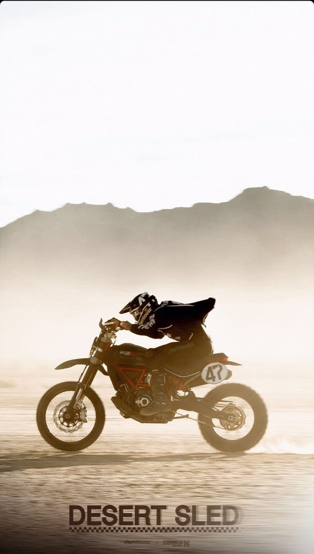 Motocross rider in full gear soaring mid-air over a dusty desert floor with hazy mountains in the background.