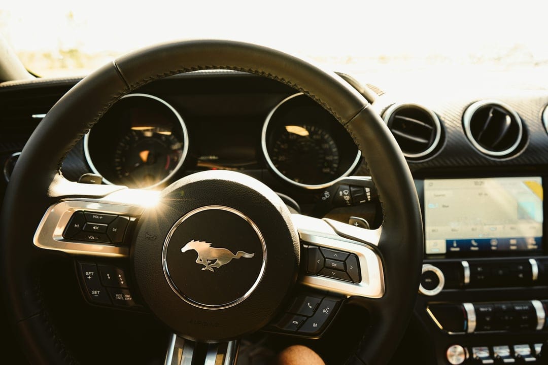 Ford Mustang steering wheel with pony logo and dashboard gauges illuminated by golden sun flare highlights.