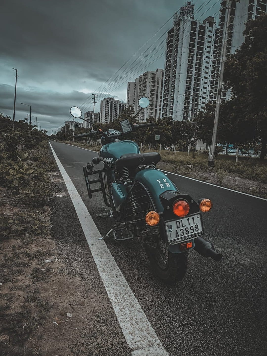 Teal Royal Enfield motorcycle parked on a city street with modern apartment buildings under a cloudy sky.