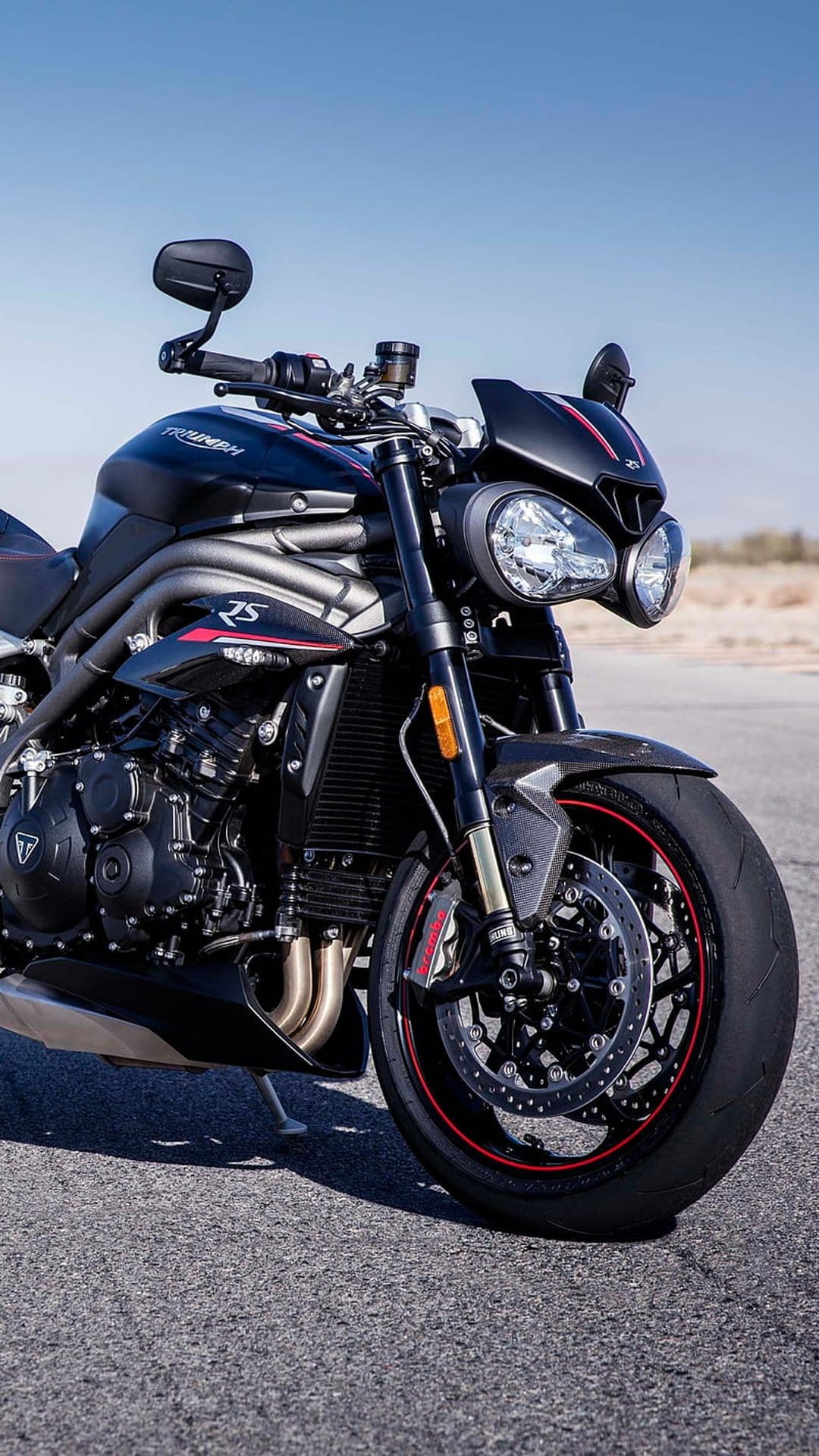 Black Triumph Speed Triple 1200 RS motorcycle with twin headlights and red-rimmed wheels parked on grey asphalt.