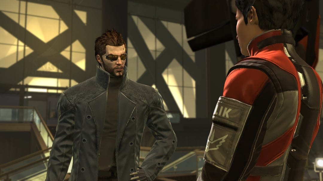 Adam Jensen wearing augmented sunglasses and a dark coat talks to an officer in a golden futuristic lobby.