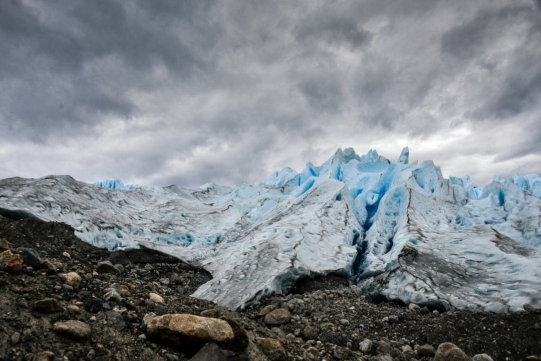 Blue glacier ice with deep crevasses sits beneath dark stormy clouds and above a rocky mountain moraine slope.