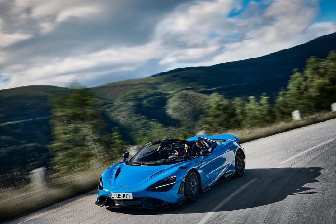 Blue convertible supercar driving fast on a winding mountain road with green hills and cloudy sky behind.
