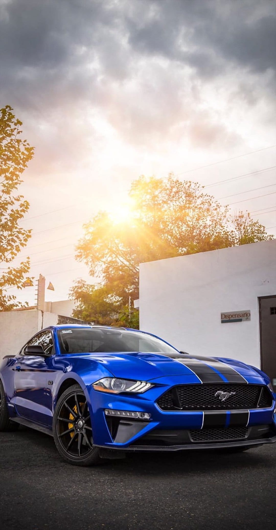 Blue Ford Mustang GT with black racing stripes parked on asphalt outdoors under a sunlit cloudy sky.