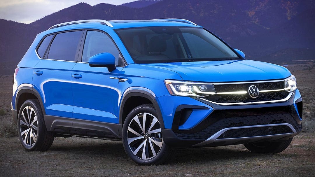 Blue Volkswagen Taos SUV with glowing headlights parked in front of misty mountains at dusk.