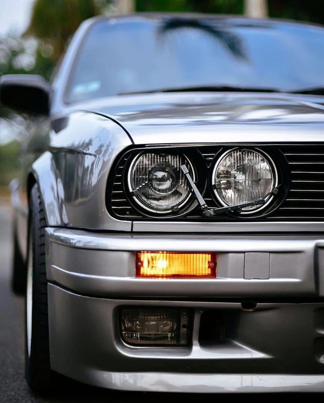 BMW E30 M3 frontal view showing silver metallic paint, dual round headlights, and a black signature kidney grille.