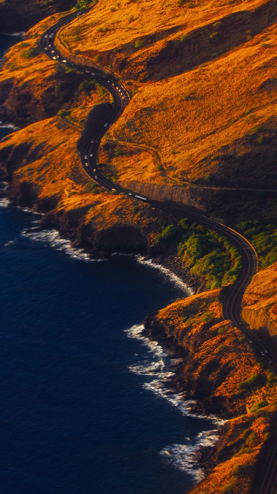 Winding asphalt road curves along golden sunlit hills beside a deep blue ocean with crashing white waves.