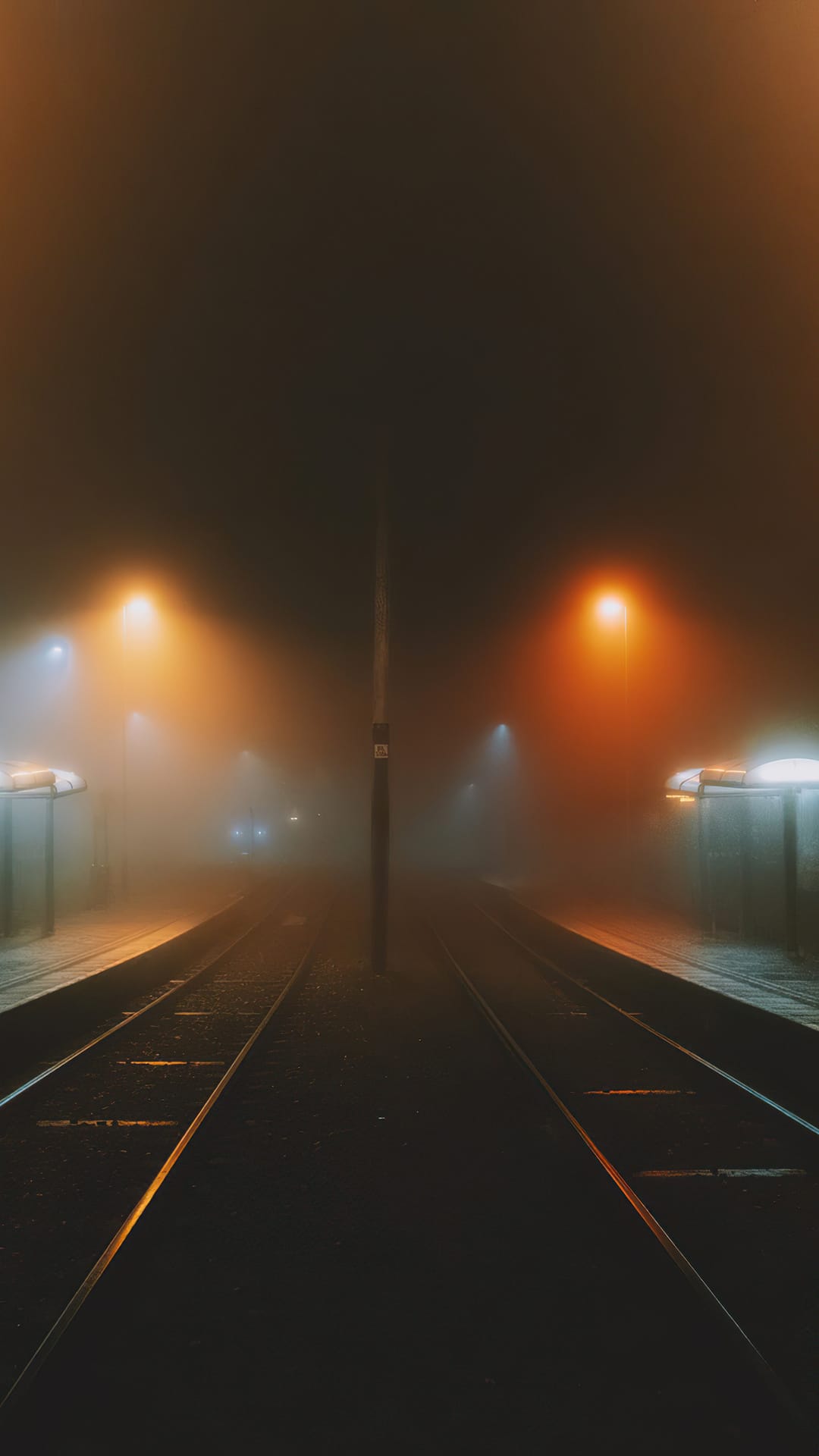 Wet railway tracks reflecting amber lights under a dense nocturnal fog, receding into an industrial horizon.
