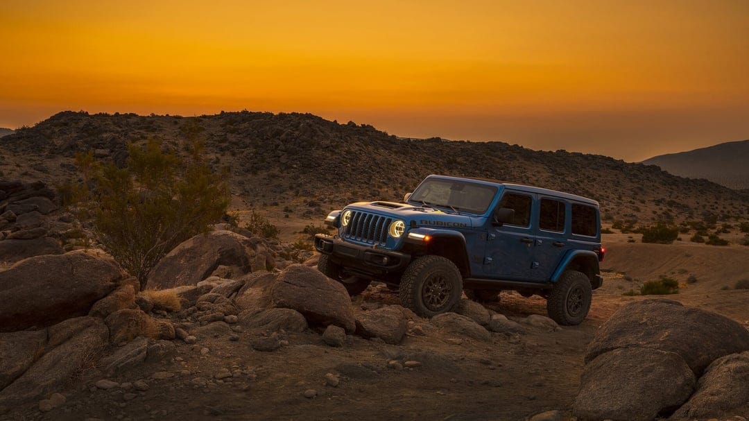 Blue Jeep Rubicon navigating a rocky desert hill during a vibrant orange sunset with glowing headlights.