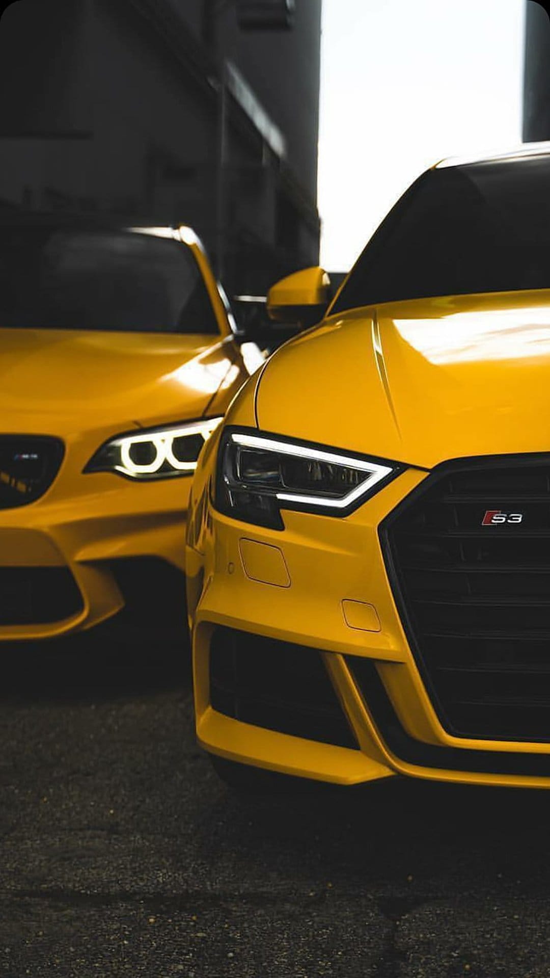 Two yellow Audi RS3 cars feature sharp LED headlights and honeycomb grilles against a dark, shadowy background.
