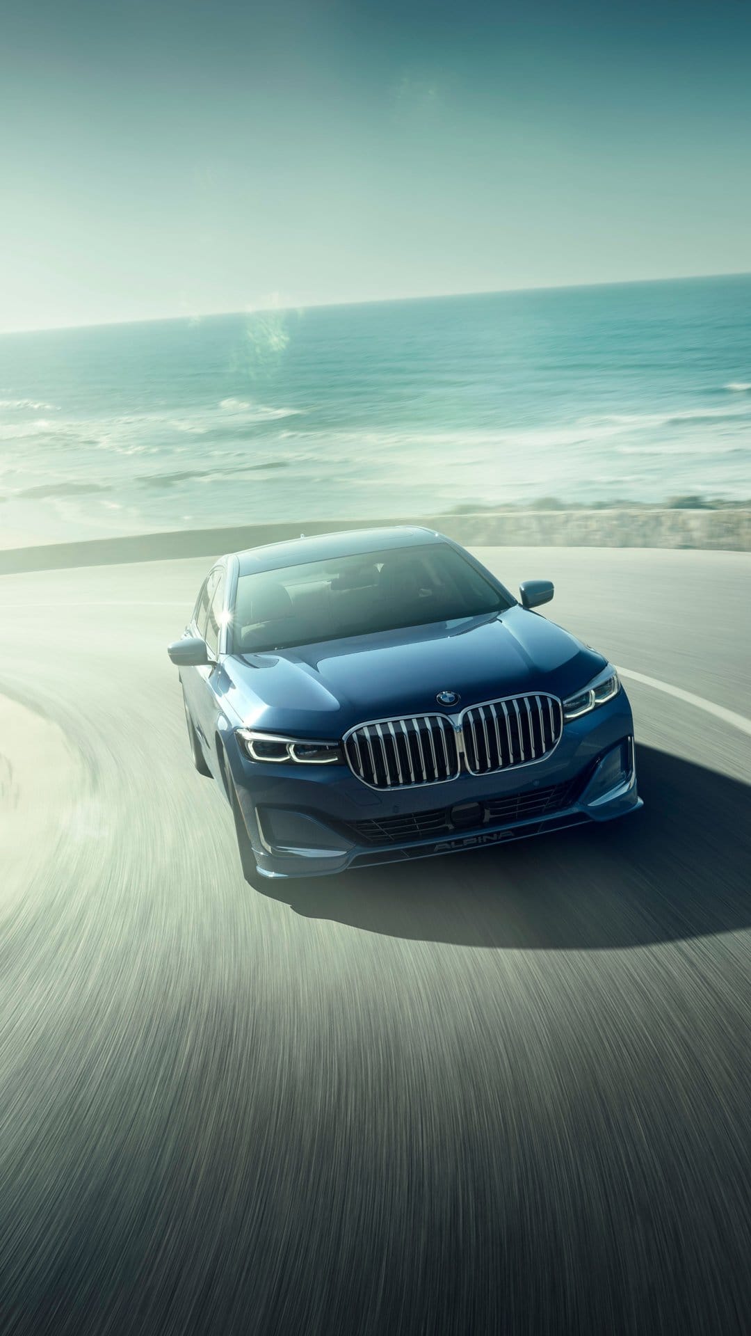 Blue Alpina sedan driving fast on a curved coastal road with ocean waves and bright sunlight flare.