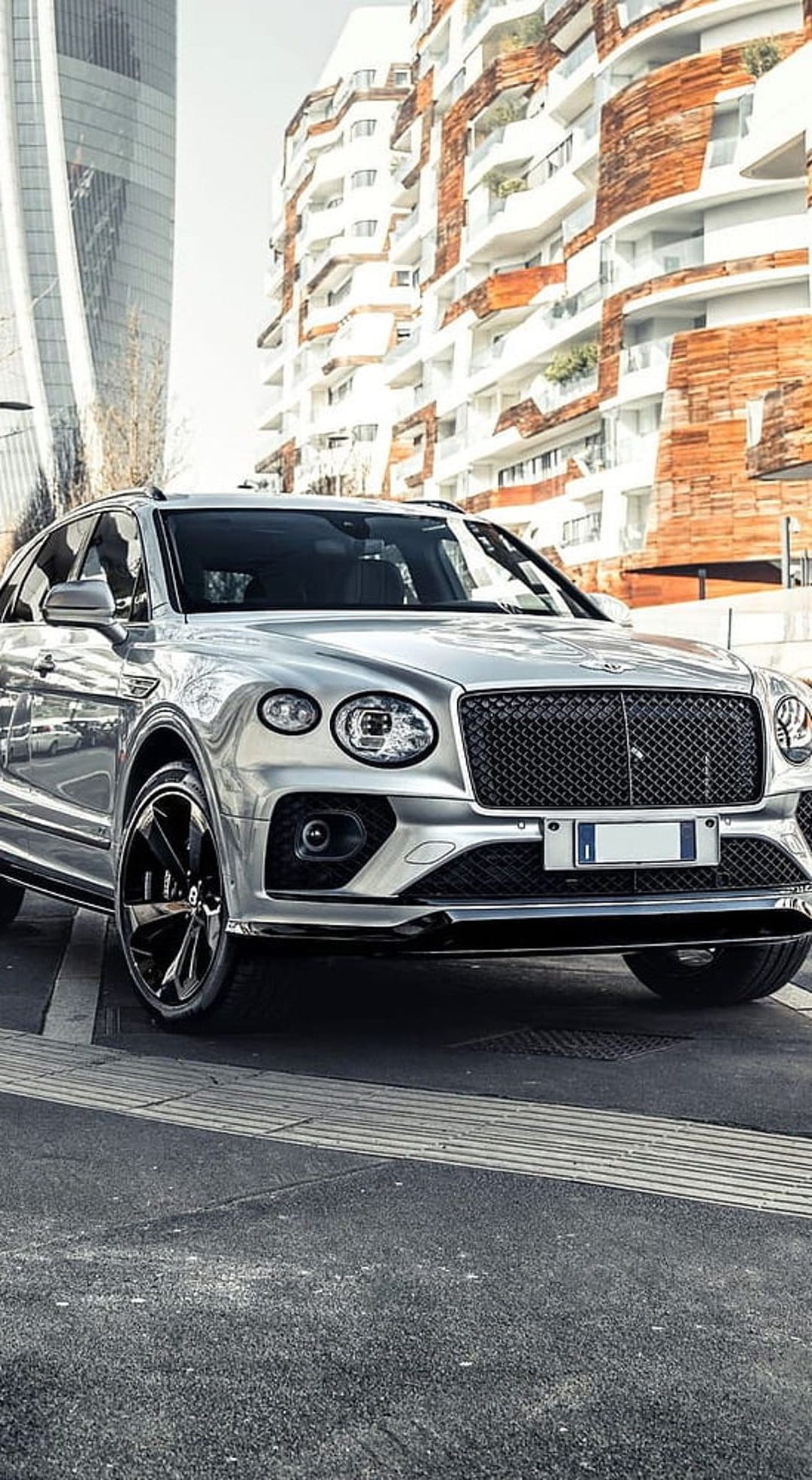 Silver Bentley Bentayga SUV parked on an urban street against a backdrop of modern geometric building facades.