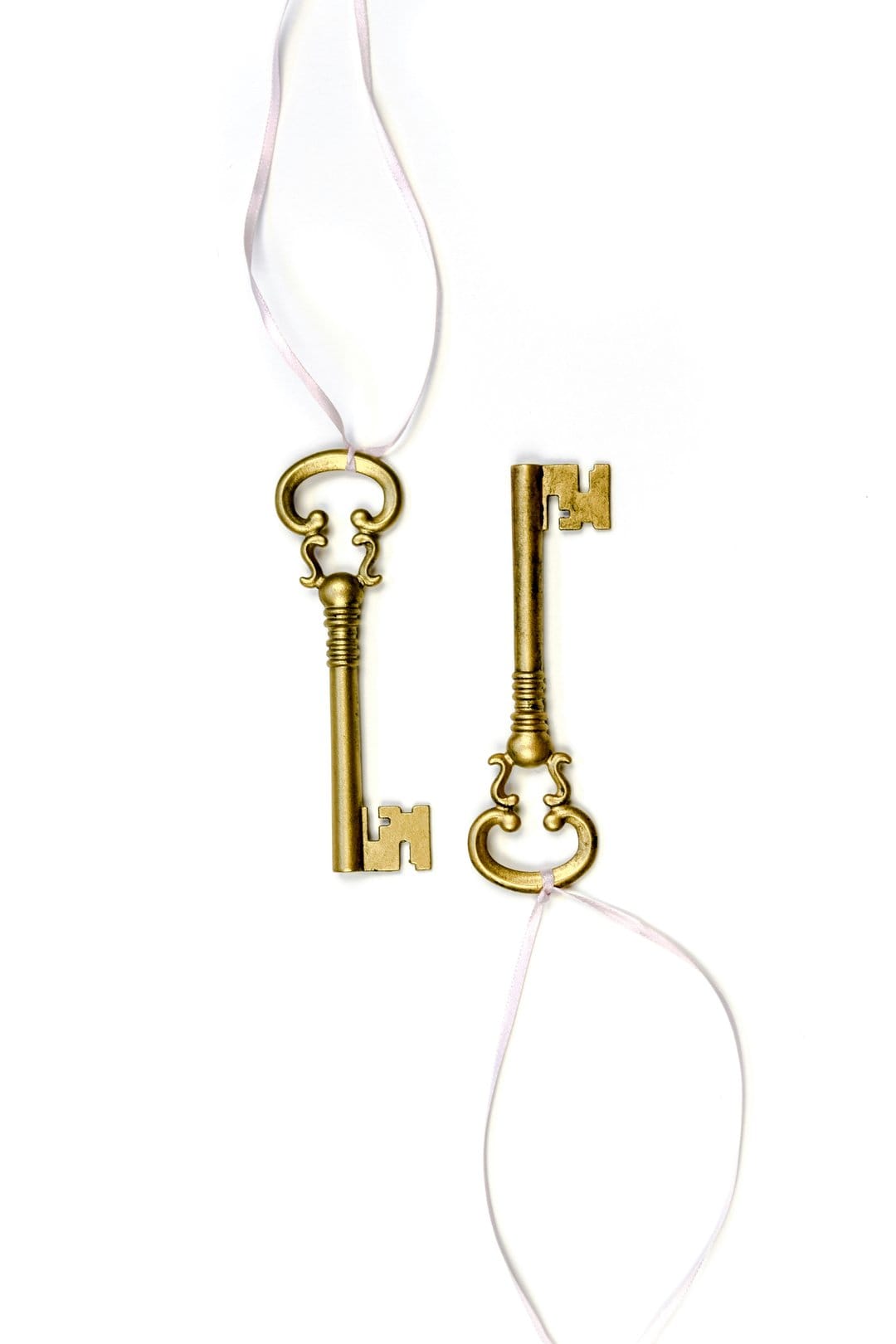 Ornate brass skeleton keys suspended by pink ribbons against a clean white background in a vertical composition.