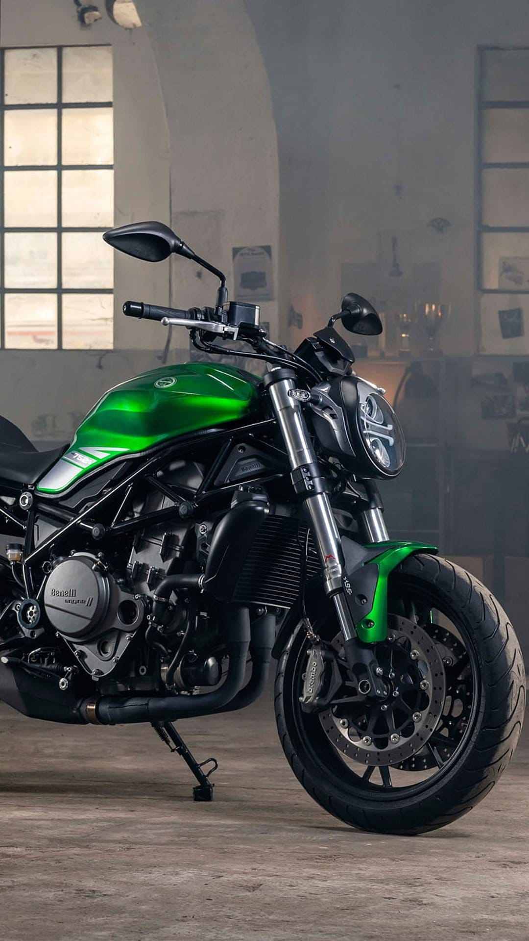 Green Benelli motorcycle with a round headlight parked inside a dimly lit industrial garage with windows.