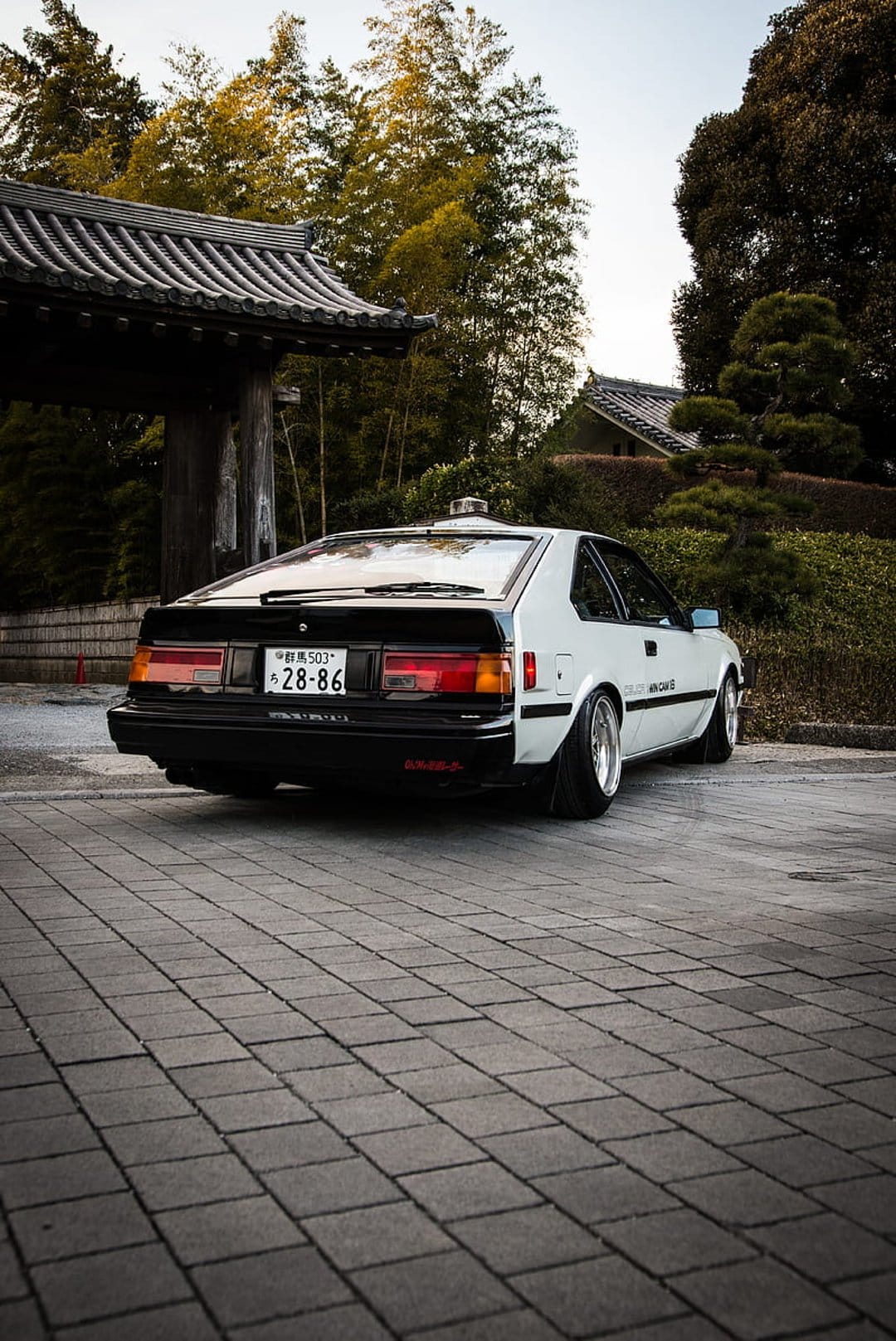 White Toyota Celica GT rear view parked on cobblestones with a traditional Japanese temple gate in the background.
