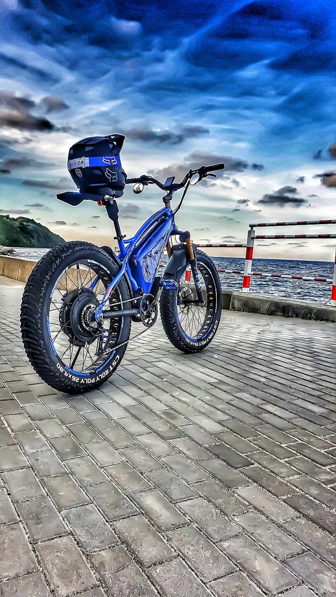 Blue fat tire electric bike parked on a brick path by the sea under a cloudy sky with a helmet on handlebars.