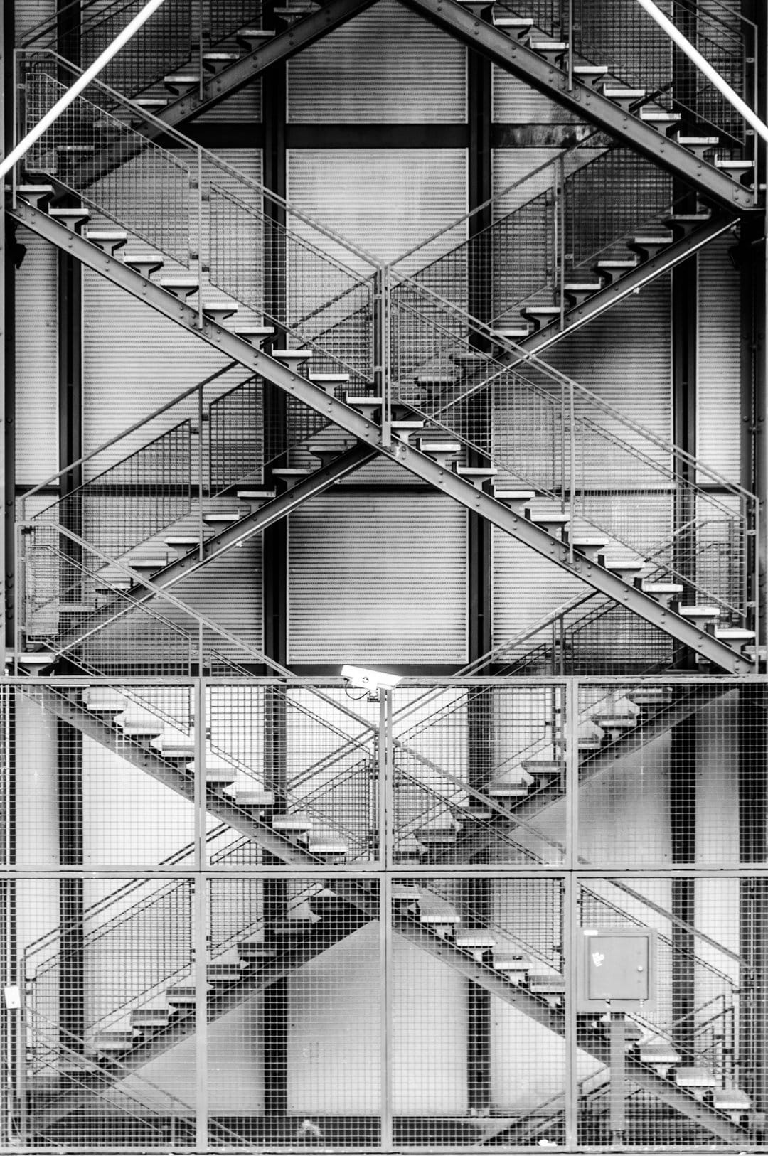 Intersecting metal staircases and wire mesh grids create a stark black and white architectural perspective.