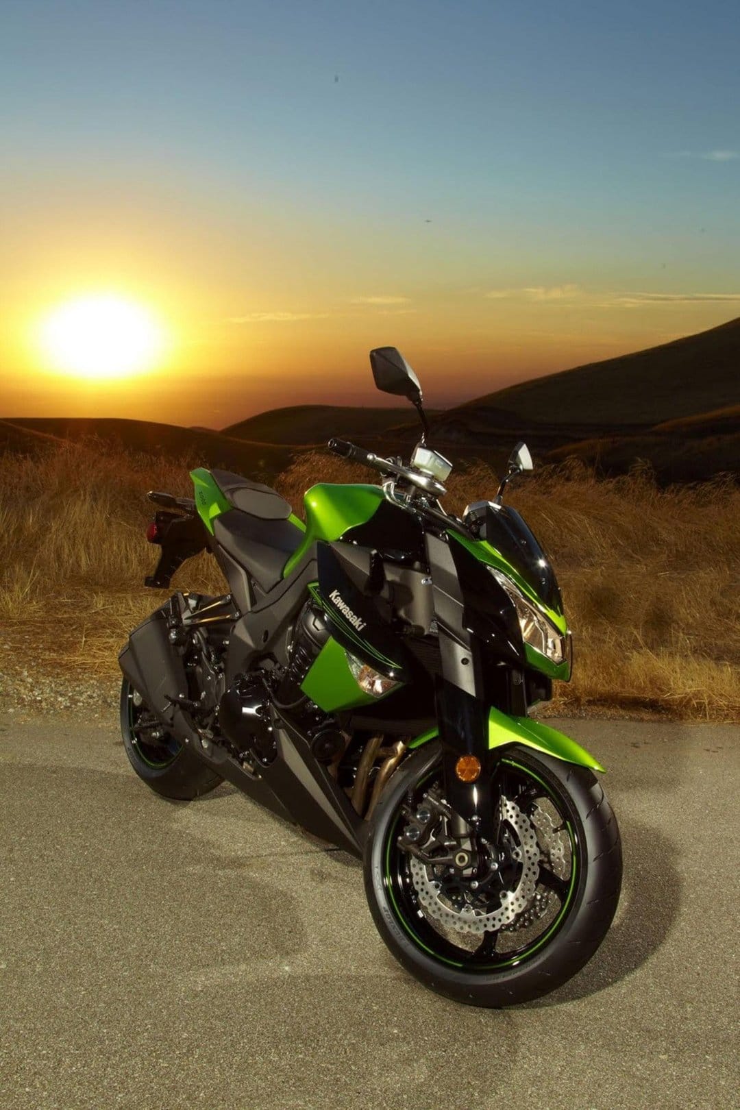 Green Kawasaki sportbike parked on an asphalt road during a golden sunset with rolling hills in the background.